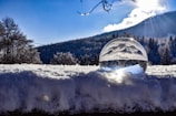 A clear crystal ball sits on a layer of fresh snow with a scenic backdrop of a snow-covered forest and mountain. Sunlight shines through the ball, reflecting and refracting light, creating an ethereal effect. The sky is clear and blue, with the sun peeking through the mountain, casting rays of light.