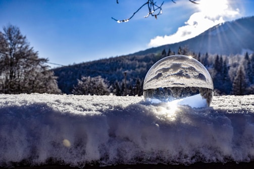 A clear crystal ball sits on a layer of fresh snow with a scenic backdrop of a snow-covered forest and mountain. Sunlight shines through the ball, reflecting and refracting light, creating an ethereal effect. The sky is clear and blue, with the sun peeking through the mountain, casting rays of light.