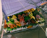 A vibrant fruit stall is filled with a variety of colorful fruits, including bananas, apples, oranges, pears, grapes, and other tropical fruits. The stall is covered by a purple tarpaulin, and several power lines are visible above it.