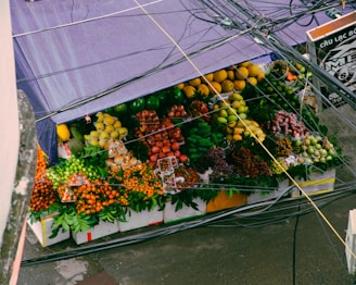 A vibrant fruit stall is filled with a variety of colorful fruits, including bananas, apples, oranges, pears, grapes, and other tropical fruits. The stall is covered by a purple tarpaulin, and several power lines are visible above it.
