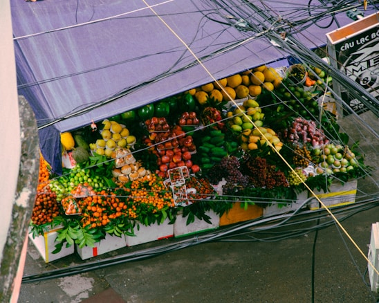 A vibrant fruit stall is filled with a variety of colorful fruits, including bananas, apples, oranges, pears, grapes, and other tropical fruits. The stall is covered by a purple tarpaulin, and several power lines are visible above it.