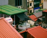 Rooftops with red and green corrugated metal sheets overlook an urban landscape. Various structures and a water tank with the word 'Tân Á' are visible. A sign for 'Hamsa Spa' can be seen in the background.