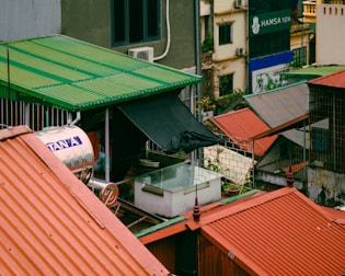 Rooftops with red and green corrugated metal sheets overlook an urban landscape. Various structures and a water tank with the word 'Tân Á' are visible. A sign for 'Hamsa Spa' can be seen in the background.