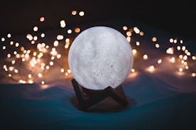 A spherical object resembling a moon lamp is positioned on a stand, surrounded by a series of soft, warm white fairy lights that create a cozy and serene ambiance. The background is dark, allowing the lights and the moon-like structure to stand out prominently.