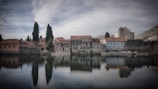 A tranquil scene of an old stone building with red-tiled roofs reflected in a calm body of water. Tall trees line the property, and a muted sky looms above. The surrounding environment exudes a sense of peacefulness and antiquity.