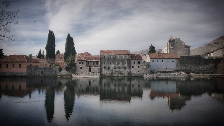 A tranquil scene of an old stone building with red-tiled roofs reflected in a calm body of water. Tall trees line the property, and a muted sky looms above. The surrounding environment exudes a sense of peacefulness and antiquity.