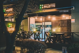A cozy-looking shop with a wooden exterior and signage that reads 'Palette'. The shop is well-lit from the inside, with simple wooden tables and chairs positioned on the front patio. The warm glow of lights creates a welcoming aura. Trees and shrubs frame the shop, enhancing its inviting atmosphere.