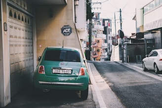 A green car is parked on the side of a narrow street adjacent to a building with a garage door. An ADT security sign is visible on the wall near the car. The street is lined with residential and commercial buildings, and there is a white car parked further down the street. Overhead, power lines cross the clear sky, and various urban architectural elements are present.