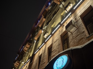 A building facade is illuminated by bright, linear lights running horizontally across its surface. The structure features a rough, textured exterior and several air conditioning units mounted on the walls. A round, glowing sign displays floral artwork near the bottom right corner.
