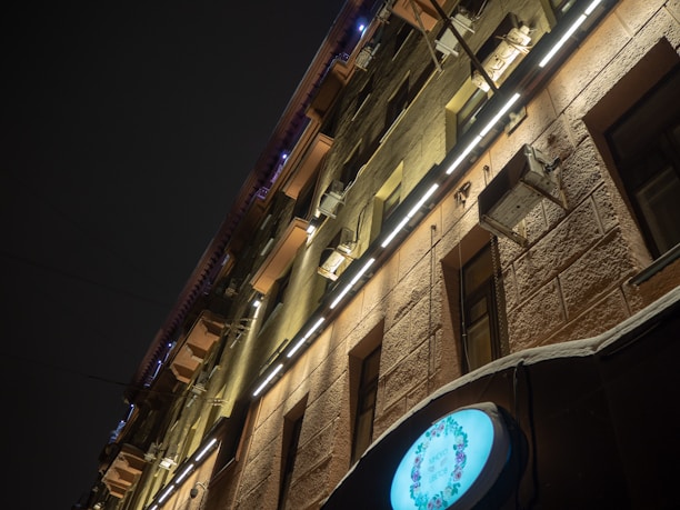 A building facade is illuminated by bright, linear lights running horizontally across its surface. The structure features a rough, textured exterior and several air conditioning units mounted on the walls. A round, glowing sign displays floral artwork near the bottom right corner.