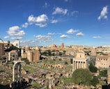 A panoramic view of ancient Roman ruins set against a backdrop of a clear blue sky with scattered white clouds. The image features historic buildings and columns, surrounded by patches of greenery. The architecture is a mix of intact and partially ruined structures, showcasing a sense of historical depth.