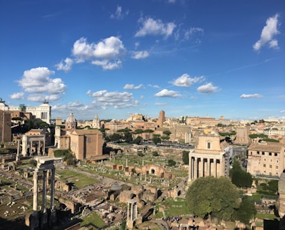 A panoramic view of ancient Roman ruins set against a backdrop of a clear blue sky with scattered white clouds. The image features historic buildings and columns, surrounded by patches of greenery. The architecture is a mix of intact and partially ruined structures, showcasing a sense of historical depth.