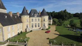 A grand historic chateau with pointed slate rooftops and a circular tower, set in a lush, manicured garden. A small red car is parked in the gravel driveway, and tall trees with rich foliage surround the building. A small pond with seating arrangements is visible to the side.