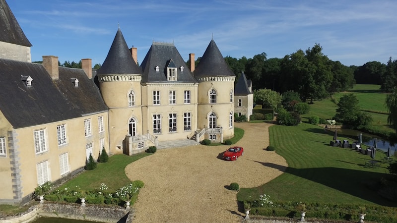 A grand historic chateau with pointed slate rooftops and a circular tower, set in a lush, manicured garden. A small red car is parked in the gravel driveway, and tall trees with rich foliage surround the building. A small pond with seating arrangements is visible to the side.