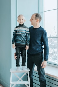 A young child stands on a white stool next to an adult who looks at the child with a smile. Both are wearing sweaters in shades of blue and black, and the interior background features a large window with natural light coming in.
