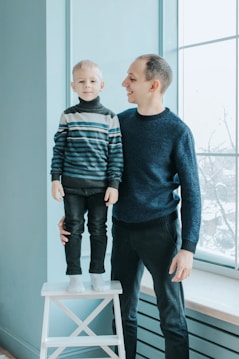 A young child stands on a white stool next to an adult who looks at the child with a smile. Both are wearing sweaters in shades of blue and black, and the interior background features a large window with natural light coming in.