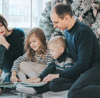 girl and boy reading book sitting between man and woman beside Christmas tree
