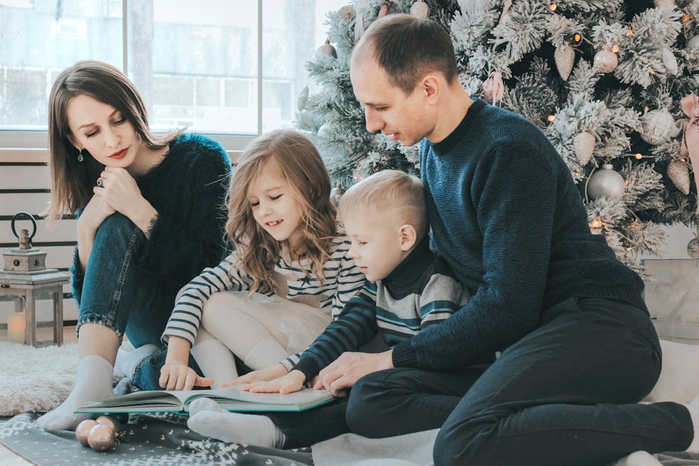 girl and boy reading book sitting between man and woman beside Christmas tree