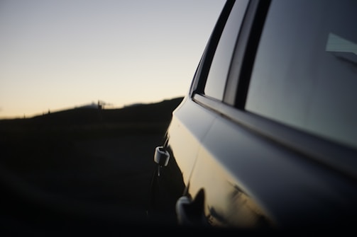 An automobile is partially visible, focusing on the door and mirror in a low-light environment. The background features a silhouette of a hilly landscape during sunset or sunrise, with a gradient sky transitioning from light to dark.