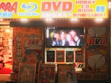 A storefront is brightly lit with an assortment of colorful posters and signs advertising DVDs, Blu-ray discs, books, and hobbies. The signs include both English and Japanese text. A TV screen displays a group of smiling people inside the store. The decor features vivid hues and animated characters, contributing to a lively and bustling atmosphere.