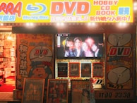 A storefront is brightly lit with an assortment of colorful posters and signs advertising DVDs, Blu-ray discs, books, and hobbies. The signs include both English and Japanese text. A TV screen displays a group of smiling people inside the store. The decor features vivid hues and animated characters, contributing to a lively and bustling atmosphere.