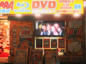 A storefront is brightly lit with an assortment of colorful posters and signs advertising DVDs, Blu-ray discs, books, and hobbies. The signs include both English and Japanese text. A TV screen displays a group of smiling people inside the store. The decor features vivid hues and animated characters, contributing to a lively and bustling atmosphere.