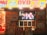 A storefront is brightly lit with an assortment of colorful posters and signs advertising DVDs, Blu-ray discs, books, and hobbies. The signs include both English and Japanese text. A TV screen displays a group of smiling people inside the store. The decor features vivid hues and animated characters, contributing to a lively and bustling atmosphere.