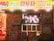 A storefront is brightly lit with an assortment of colorful posters and signs advertising DVDs, Blu-ray discs, books, and hobbies. The signs include both English and Japanese text. A TV screen displays a group of smiling people inside the store. The decor features vivid hues and animated characters, contributing to a lively and bustling atmosphere.