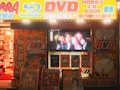 A storefront is brightly lit with an assortment of colorful posters and signs advertising DVDs, Blu-ray discs, books, and hobbies. The signs include both English and Japanese text. A TV screen displays a group of smiling people inside the store. The decor features vivid hues and animated characters, contributing to a lively and bustling atmosphere.