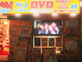 A storefront is brightly lit with an assortment of colorful posters and signs advertising DVDs, Blu-ray discs, books, and hobbies. The signs include both English and Japanese text. A TV screen displays a group of smiling people inside the store. The decor features vivid hues and animated characters, contributing to a lively and bustling atmosphere.
