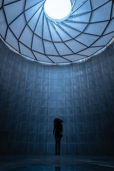 A figure stands centrally in a futuristic, cylindrical space with a geometric, spiral-patterned ceiling that opens to a bright, circular light source. The environment is composed of smooth, metallic panels with a cool blue tone, creating a surreal and dramatic atmosphere.