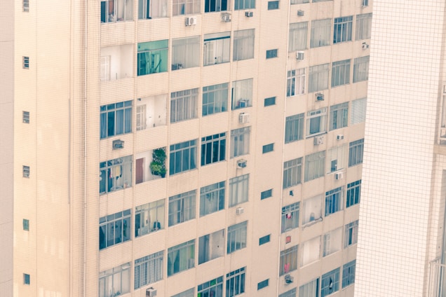 A high-rise apartment building with numerous windows, some of which have open curtains or blinds. The exterior is made of light-colored bricks, and air conditioning units are attached beneath several windows. A small balcony with green plants can be seen among the windows.