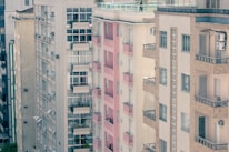 A view of several residential apartment buildings closely aligned. The architecture is characterized by large windows, small balconies, and air conditioning units visible on the facade. The buildings are primarily neutral tones with one featuring prominent pink accents on the balconies.