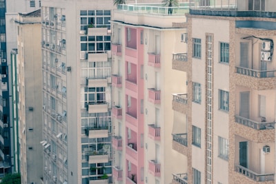 A view of several residential apartment buildings closely aligned. The architecture is characterized by large windows, small balconies, and air conditioning units visible on the facade. The buildings are primarily neutral tones with one featuring prominent pink accents on the balconies.