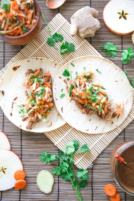 Two tortillas filled with shredded meat and topped with a colorful slaw made from carrots and herbs. Surrounding the tortillas are items such as ginger, apple slices, carrot slices, and a small bowl containing more of the slaw. Cilantro leaves are scattered across a bamboo mat, and a cutting board along with a bowl of sauce can be seen.