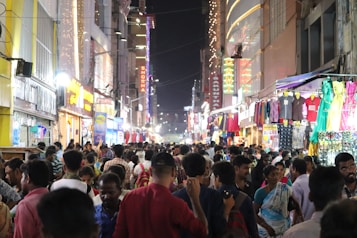 A bustling night market scene with a crowded street, lined with brightly lit shops on both sides. Numerous people are shopping, walking, and interacting with street vendors. The vibrant storefronts display colorful clothing and other goods, creating a lively and energetic atmosphere.