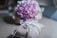 A decorative burlap pillow with lace and ribbons holds a pair of metallic rings. In the blurred background, a bouquet of pink and purple flowers is visible, creating a soft, romantic atmosphere.