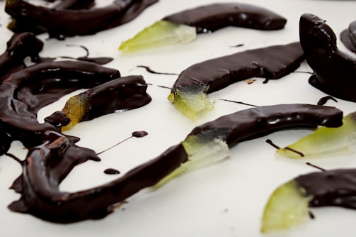 Slices of fruit, possibly citrus, are coated in dark chocolate and arranged on a white surface. The fruit appears translucent with a greenish hue, providing contrast to the glossy, dark coating.