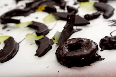 A close-up view of various sliced fruits and a donut covered in glossy dark chocolate, arranged on a white surface. The fruits appear to include some green slices, which could be apples or pears.