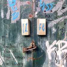 A metal door covered in peeling paint, graffiti, and writings in various colors, including blue and white. Two metal plates are mounted on the door with the letters 'Li' and 'Fe' written in blue. A rusted padlock secures the door.