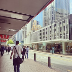 A bustling urban street scene featuring several people walking along a sidewalk next to tall, modern office buildings with reflective glass windows. Retail stores line the street with visible signage, and the atmosphere appears lively and busy.