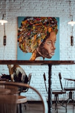 A vibrant painting of a woman's face with an abstract, colorful headwrap hanging on a white brick wall. The artwork combines earthy and bright tones with splatters and layers creating a textured effect. Below the painting, a cafe setting features a person with long hair sitting at a table, immersed in writing or drawing. The room features wooden chairs, a table, and warm lighting fixtures hanging on both sides.