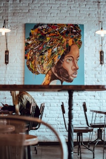 A vibrant painting of a woman's face with an abstract, colorful headwrap hanging on a white brick wall. The artwork combines earthy and bright tones with splatters and layers creating a textured effect. Below the painting, a cafe setting features a person with long hair sitting at a table, immersed in writing or drawing. The room features wooden chairs, a table, and warm lighting fixtures hanging on both sides.