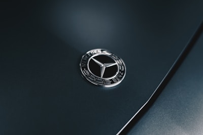 A close-up of a Mercedes-Benz emblem on a dark-colored car surface. The emblem features a silver three-pointed star encircled with the brand name in a sleek, metallic finish.