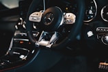A close-up view of a luxury car interior featuring a Mercedes-Benz steering wheel with AMG branding. The dashboard displays high-quality materials, modern design, and advanced control buttons.