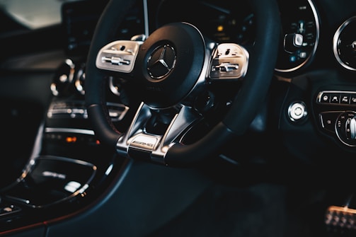 A close-up view of a luxury car interior featuring a Mercedes-Benz steering wheel with AMG branding. The dashboard displays high-quality materials, modern design, and advanced control buttons.