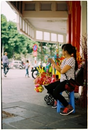 A person is seated on a small blue plastic stool on a sidewalk, holding colorful water guns. Various toys, including balloons and other plastic items, are displayed on a cart beside them. Trees and a street can be seen in the background, with people and vehicles moving by.