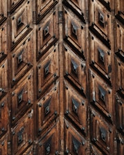 Intricately carved wooden surface featuring a pattern of repeated geometric shapes, with diamond-like designs and a rich, natural wood texture.