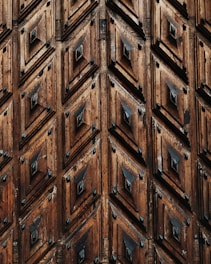 Intricately carved wooden surface featuring a pattern of repeated geometric shapes, with diamond-like designs and a rich, natural wood texture.