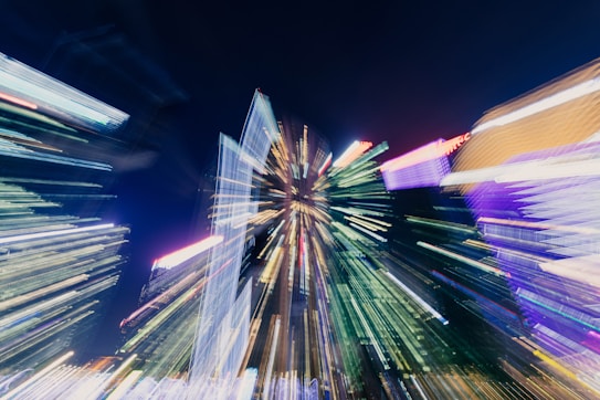 A cityscape at night with lights creating a dynamic, motion-blurred effect, radiating from a central point. The vivid colors and streaks suggest movement and energy, emphasizing the hustle and bustle of urban life.